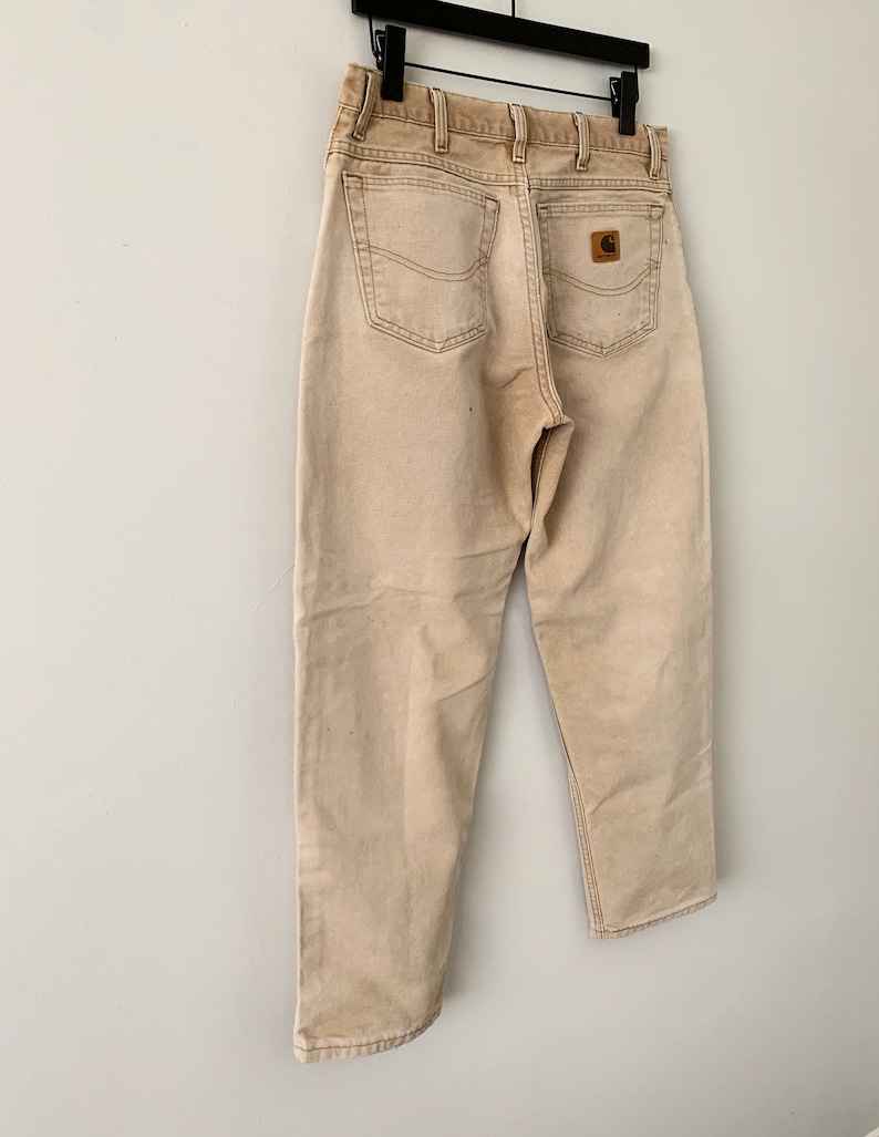Puede incluir: Un par de pantalones vaqueros Carhartt de color marr&oacute;n claro con un parche de cuero marr&oacute;n en el bolsillo trasero.