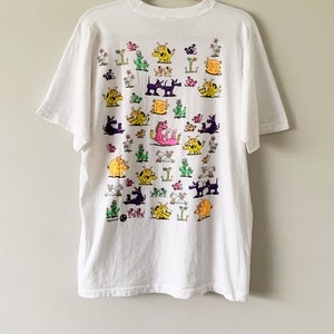 May include: White t-shirt with a colorful cartoon print of various animals, including dogs, cats, and dragons. The print is all over the shirt and features a variety of colors, including pink, yellow, green, and purple.