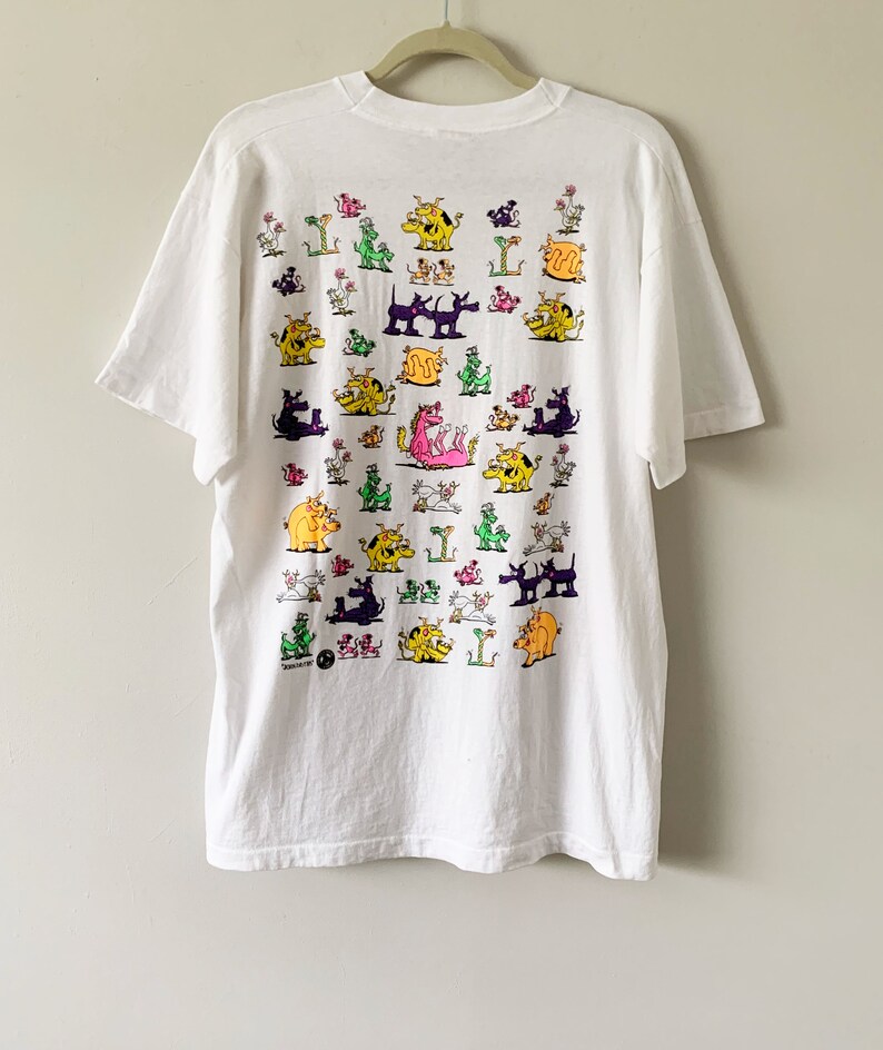 May include: White t-shirt with a colorful cartoon print of various animals, including dogs, cats, and dragons. The print is all over the shirt and features a variety of colors, including pink, yellow, green, and purple.