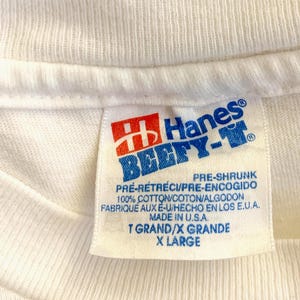 May include: Close-up of a white Hanes Beefy-T shirt tag. The tag features the Hanes logo in red and blue, with text indicating it is pre-shrunk and made of 100% cotton. The size is X-Large.