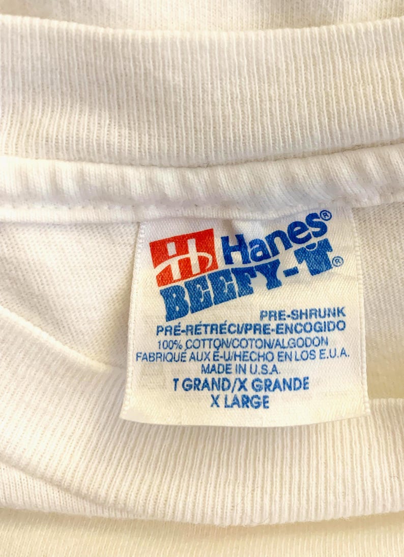 May include: Close-up of a white Hanes Beefy-T shirt tag. The tag features the Hanes logo in red and blue, with text indicating it is pre-shrunk and made of 100% cotton. The size is X-Large.