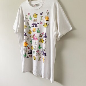 May include: White t-shirt with a colorful cartoon print of various animals. The animals are all different colors and have different expressions. The shirt is a vintage style and has a crew neck.