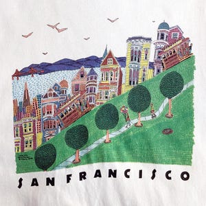 May include: White t-shirt featuring a colorful illustration of San Francisco. The design includes cable cars, Victorian houses, a green hillside, and the text "SAN FRANCISCO" in black letters. The image also includes a few birds in the sky.