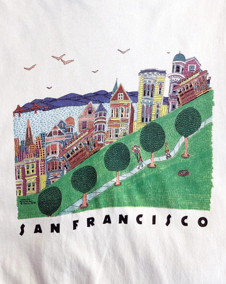 May include: White t-shirt featuring a colorful illustration of San Francisco. The design includes cable cars, Victorian houses, a green hillside, and the text "SAN FRANCISCO" in black letters. The image also includes a few birds in the sky.