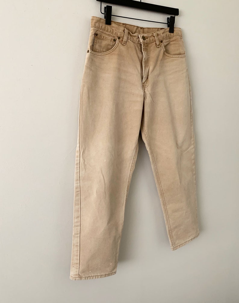 Puede incluir: Un par de pantalones vaqueros de color marr&oacute;n claro colgados de una percha. Los vaqueros tienen una pierna recta y un ajuste holgado.