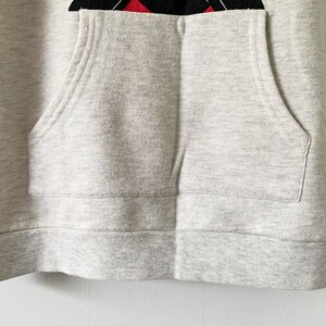 Puede incluir: Una sudadera con capucha gris con un bolsillo en la parte delantera. La sudadera tiene un dobladillo y pu&ntilde;os de canal&eacute;.