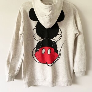 Puede incluir: Una sudadera gris con capucha con un gr&aacute;fico de Mickey Mouse negro y rojo en la espalda. El gr&aacute;fico muestra a Mickey Mouse sentado de espaldas al espectador, con sus caracter&iacute;sticos pantalones cortos rojos y dos c&iacute;rculos negros en sus pantalones cortos.