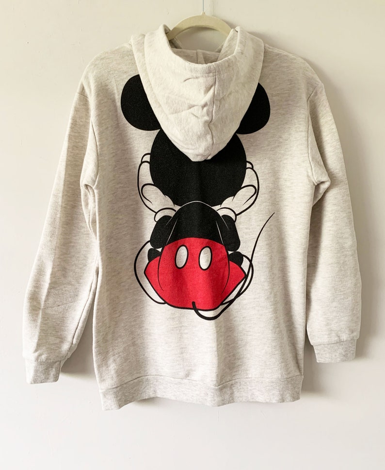 Puede incluir: Una sudadera gris con capucha con un gr&aacute;fico de Mickey Mouse negro y rojo en la espalda. El gr&aacute;fico muestra a Mickey Mouse sentado de espaldas al espectador, con sus caracter&iacute;sticos pantalones cortos rojos y dos c&iacute;rculos negros en sus pantalones cortos.