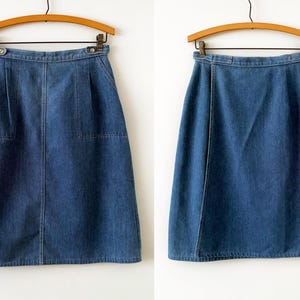 May include: A medium-wash blue denim skirt featuring a high-waisted design with a button closure at the waistband, two front pockets, and a straight-cut silhouette. The skirt is displayed on a wooden hanger, showing both the front and back views.