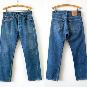 Puede incluir: Un par de pantalones vaqueros de mezclilla azul mostrados de frente y de espaldas, colgados en perchas de madera. Los vaqueros cuentan con cierre de botones, cinco bolsillos, remaches color cobre y una etiqueta de cuero beige con texto.