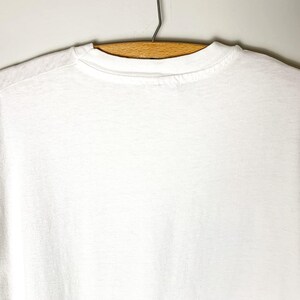 May include: A plain white t-shirt hangs on a wooden hanger against a light-colored wall. The t-shirt is made of a soft, opaque fabric. The neckline and shoulders are visible, showing the garment's simple design.