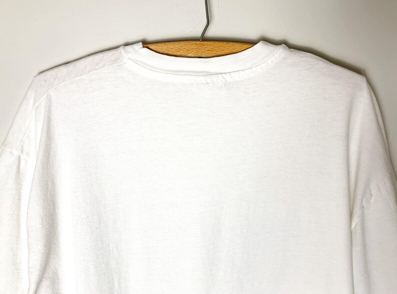 May include: A plain white t-shirt hangs on a wooden hanger against a light-colored wall. The t-shirt is made of a soft, opaque fabric. The neckline and shoulders are visible, showing the garment's simple design.