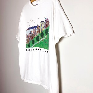 May include: White t-shirt featuring a colorful illustration of San Francisco's iconic landmarks, including Victorian houses and a cable car, with the city's name printed below. The shirt is hanging on a wooden hanger.