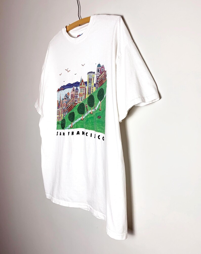 May include: White t-shirt featuring a colorful illustration of San Francisco's iconic landmarks, including Victorian houses and a cable car, with the city's name printed below. The shirt is hanging on a wooden hanger.