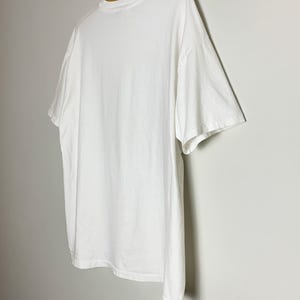 May include: A plain white t-shirt hangs on a wooden hanger against a light gray wall. The t-shirt is made of a soft, opaque material and has a crew neck and short sleeves. The shirt is slightly oversized.