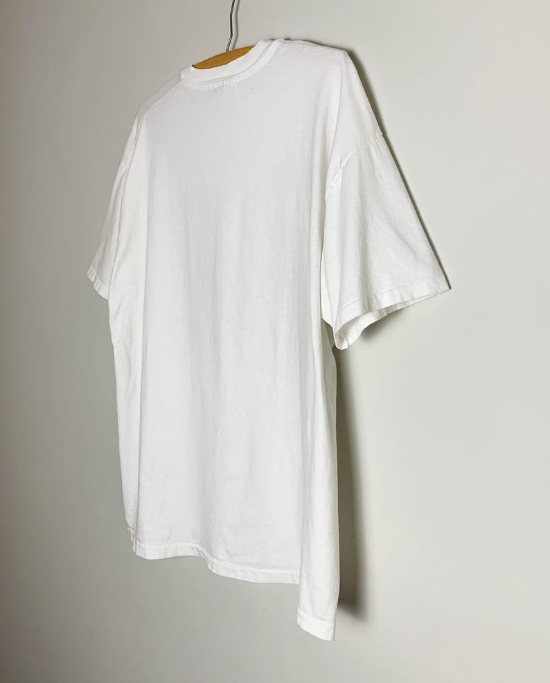 May include: A plain white t-shirt hangs on a wooden hanger against a light gray wall. The t-shirt is made of a soft, opaque material and has a crew neck and short sleeves. The shirt is slightly oversized.