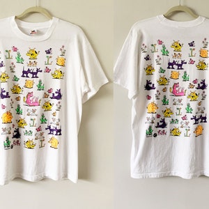 May include: White t-shirt with a colorful graphic print of various cartoon animals. The print features a variety of animals, including dogs, cats, and dragons, all in different colors and poses.