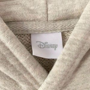 Puede incluir: Primer plano de una sudadera con capucha gris claro con una etiqueta blanca que dice "Disney".