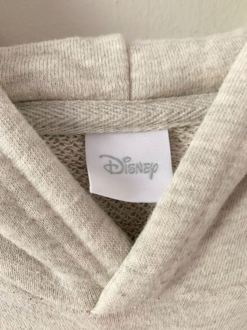 Puede incluir: Primer plano de una sudadera con capucha gris claro con una etiqueta blanca que dice "Disney".