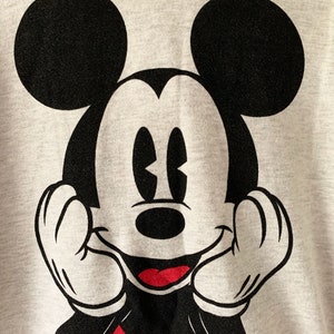 Puede incluir: Una sudadera gris con una imagen en blanco y negro de Mickey Mouse. Mickey est&aacute; sonriendo y tiene las manos levantadas hacia sus mejillas.