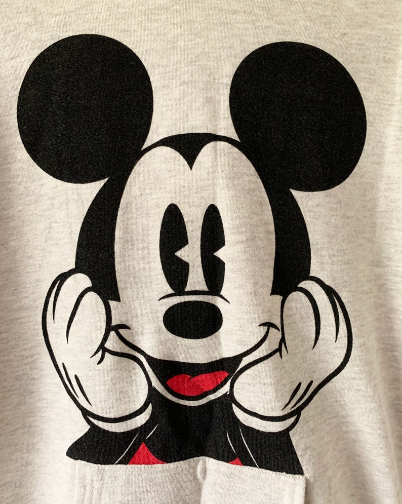 Puede incluir: Una sudadera gris con una imagen en blanco y negro de Mickey Mouse. Mickey est&aacute; sonriendo y tiene las manos levantadas hacia sus mejillas.