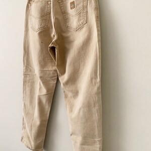 Puede incluir: Un par de pantalones vaqueros Carhartt de color beige colgados de una percha. Los vaqueros tienen un aspecto descolorido y est&aacute;n hechos de una mezcla de algod&oacute;n resistente.