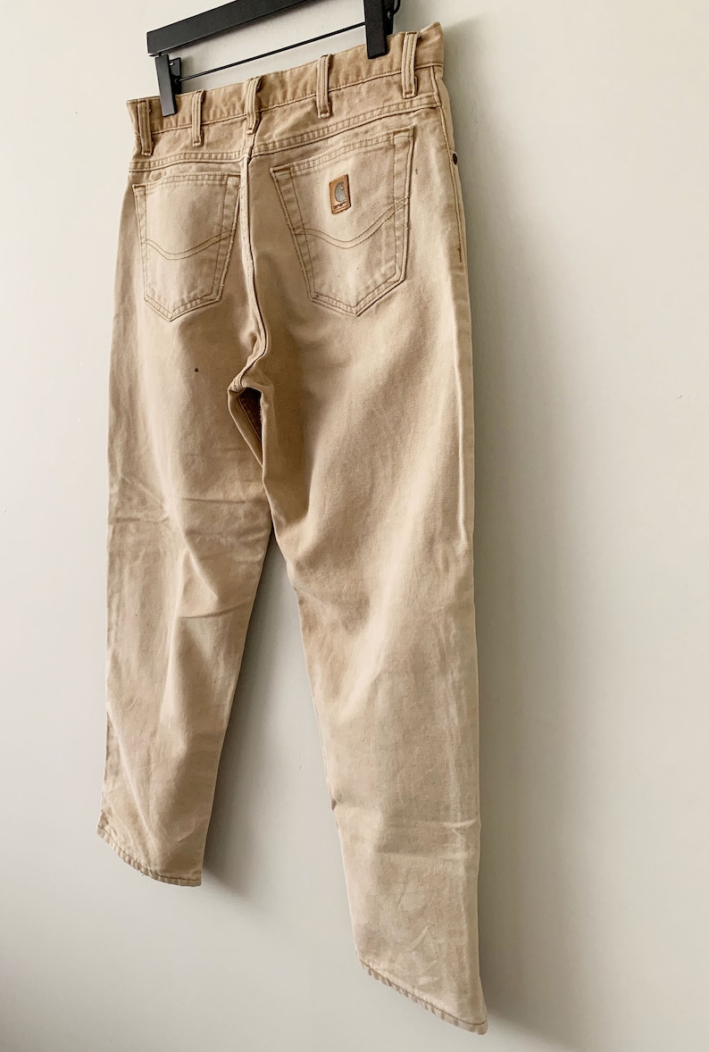 Puede incluir: Un par de pantalones vaqueros Carhartt de color beige colgados de una percha. Los vaqueros tienen un aspecto descolorido y est&aacute;n hechos de una mezcla de algod&oacute;n resistente.