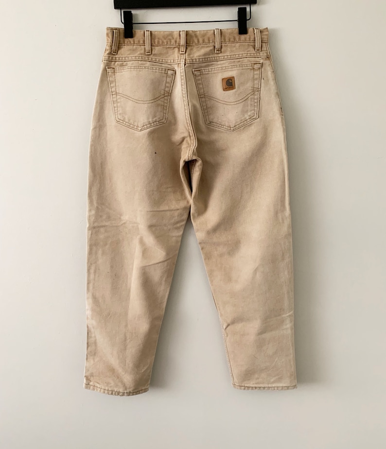 Puede incluir: Un par de pantalones vaqueros Carhartt de color marr&oacute;n claro con un parche de cuero marr&oacute;n en el bolsillo trasero.