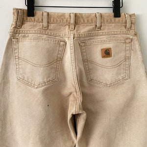 Puede incluir: Un par de pantalones vaqueros de color marr&oacute;n claro con dos bolsillos traseros y un parche con el logotipo de Carhartt en el bolsillo trasero derecho.