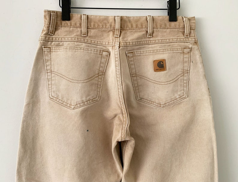 Puede incluir: Un par de pantalones vaqueros de color marr&oacute;n claro con dos bolsillos traseros y un parche con el logotipo de Carhartt en el bolsillo trasero derecho.