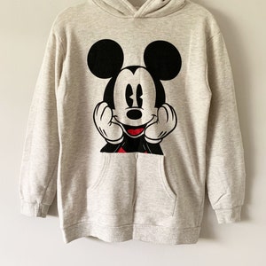 Puede incluir: Una sudadera con capucha de color gris claro con un gr&aacute;fico de Mickey Mouse en negro y rojo en la parte delantera. El gr&aacute;fico muestra a Mickey Mouse con las manos en las mejillas y una gran sonrisa.
