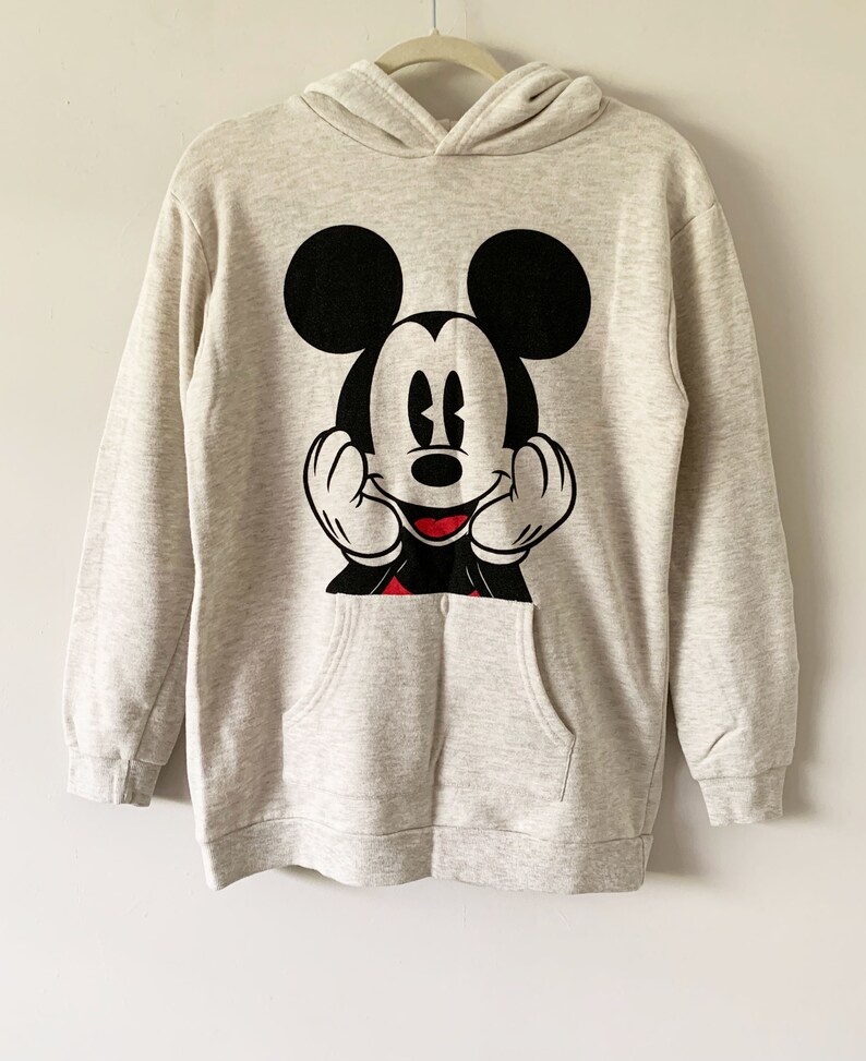 Puede incluir: Una sudadera con capucha de color gris claro con un gr&aacute;fico de Mickey Mouse en negro y rojo en la parte delantera. El gr&aacute;fico muestra a Mickey Mouse con las manos en las mejillas y una gran sonrisa.