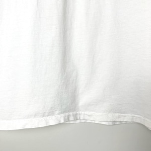 May include: Close-up of a white t-shirt. The fabric has a slightly textured appearance with subtle vertical folds. The bottom hem is visible, showing a folded and stitched edge.
