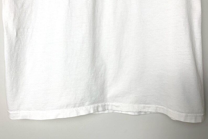 May include: Close-up of a white t-shirt. The fabric has a slightly textured appearance with subtle vertical folds. The bottom hem is visible, showing a folded and stitched edge.