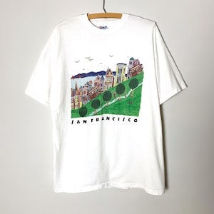 May include: White t-shirt featuring a colorful illustration of San Francisco's iconic cityscape, including Victorian houses, a cable car, and the text "SAN FRANCISCO" in black. The design is set against a green hillside.
