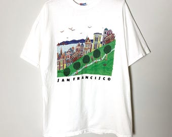 May include: White t-shirt featuring a colorful illustration of San Francisco's iconic cityscape, including Victorian houses, a cable car, and the text "SAN FRANCISCO" in black. The design is set against a green hillside.