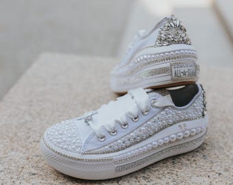 May include: White sneakers with a pearl and rhinestone embellishment. The sneakers have a platform sole and a white satin ribbon tied around the ankle. The brand name "Converse All-Star" is visible on the side of the shoe.