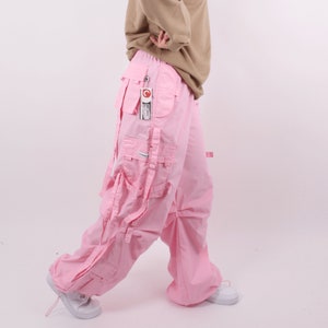 Peut inclure: Pantalon cargo rose avec de multiples poches et sangles. Le pantalon est ample et fabriqué dans un tissu léger.