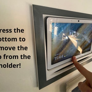 Op de afbeelding: Een witte tablet met een grijs frame is gemonteerd aan een muur. De tablet toont een klok met de tijd 11:44. Een vinger wijst naar de onderkant van de tablet, wat aangeeft dat de tablet kan worden verwijderd door op de onderkant te drukken.