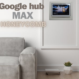 Peut inclure: Une image encadrée blanche avec un écran intelligent Google Hub Max Honeycomb à l'intérieur. L'écran affiche une image d'une scène de plage avec l'heure 13:03. L'image est accrochée à un mur blanc à côté d'un canapé gris.