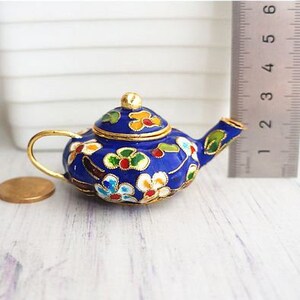 May include: A small blue teapot with a gold lid and floral designs in shades of red, yellow, and white. The teapot is made of enamel and has a delicate, ornate design.