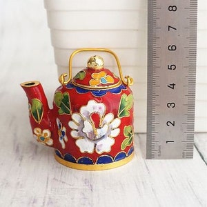 May include: A small red teapot with a gold handle and spout. The teapot is decorated with white and gold flowers and green leaves. The teapot is about 3 inches tall.