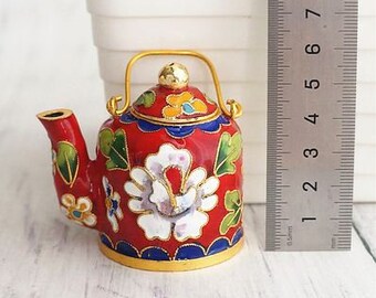 May include: A small red teapot with a gold handle and spout. The teapot is decorated with white and gold flowers and green leaves. The teapot is about 3 inches tall.
