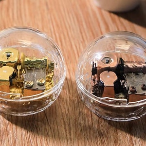 May include: Two clear glass globe music boxes with intricate gold and bronze coloured mechanisms inside. The globes are sitting on a wooden surface.