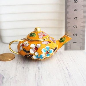 May include: A small, yellow teapot with a floral design. The teapot is decorated with pink, blue, green, and white flowers. The teapot has a gold handle and spout.