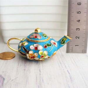 May include: A small, blue teapot with a floral design. The teapot is made of metal and has a gold trim. The flowers are pink, white, and green.