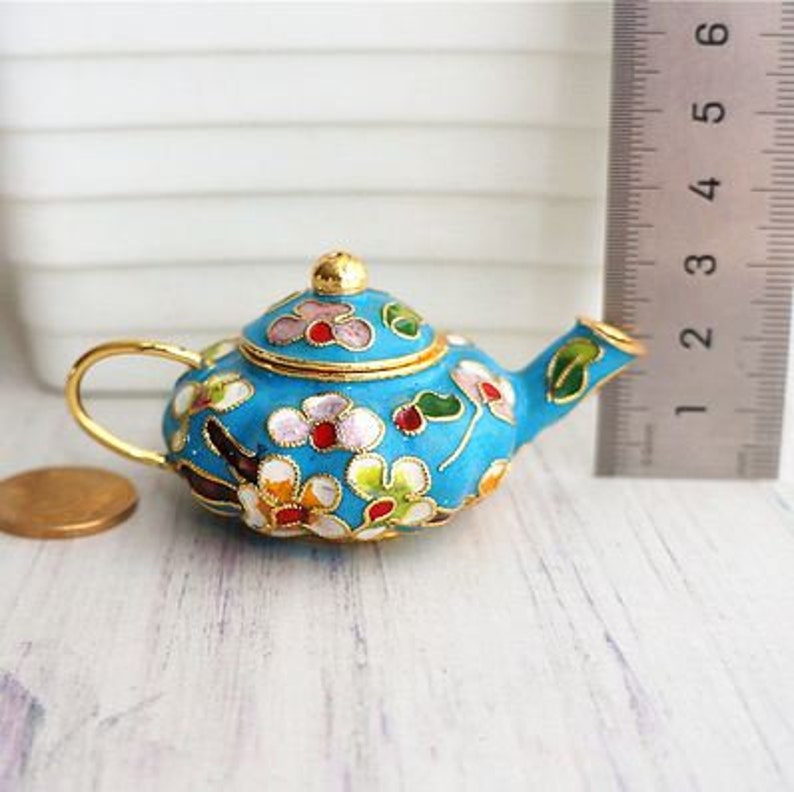 May include: A small, blue teapot with a floral design. The teapot is made of metal and has a gold trim. The flowers are pink, white, and green.
