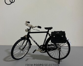 May include: A black bicycle with a black leather bag on the back rack. The bag has the letters "JY" on it. The bicycle is on a white stand. The text "FROM POOPO SELECTION ALL INTERESTING SOULS ARE WORTH HAVING" is below the bicycle.