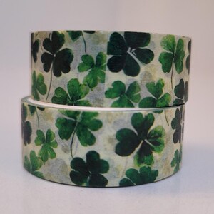 May include: Two rolls of decorative washi tape featuring a repeating pattern of green shamrocks on a light beige background. The tape is stacked, showing the detailed clover design, perfect for crafts and scrapbooking.