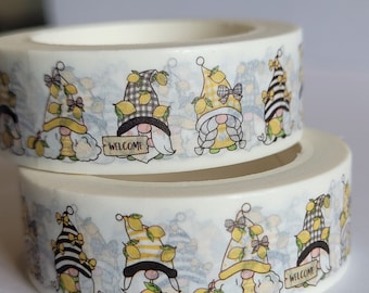 May include: Two rolls of white washi tape featuring a repeating pattern of gnomes wearing hats decorated with yellow lemons, bows, and stripes or plaid designs. Some gnomes hold a sign that reads WELCOME in black capital letters.